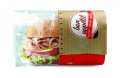 Opakowanie Snack Bag M NATURE Fifty Fifty w przezroczystym folii, z nadrukiem bon appétit i ENJOY YOUR MEAL. W środku widoczny kanapka z szynką, serem, sałatą i pomidorem. Ułożone z boku, z czerwonym dolnym paskiem.
