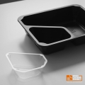 Wkładka INS-40 METROOPACK w kolorze białym, wykonana z przezroczystego plastiku, znajduje się obok czarnej, prostokątnej podstawy. Wkładka ma nieregularny kształt z wycięciem, idealna do podziału porcji. Tło jest jednolite, co uwydatnia produkt.
